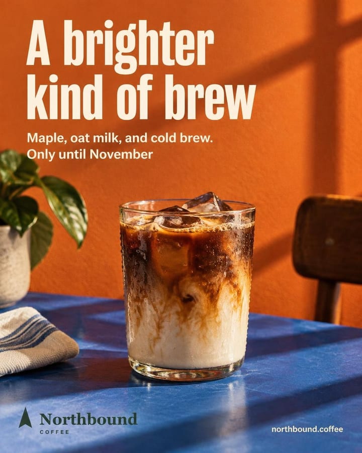 A glass of maple oat cold brew with ice and condensation sits on a cobalt blue table against a tangerine wall, crossed by long morning window shadows.